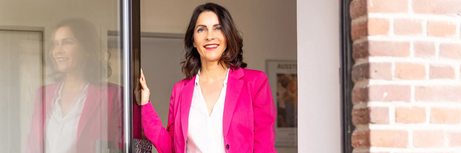 Kathrin Ipse - Expertin für Stressregulation, Nervensystem-Balance und professionelle Hypnose - Eine lächelnde Frau steht in einem Türrahmen und trägt einen leuchtend pinkfarbenen Blazer.