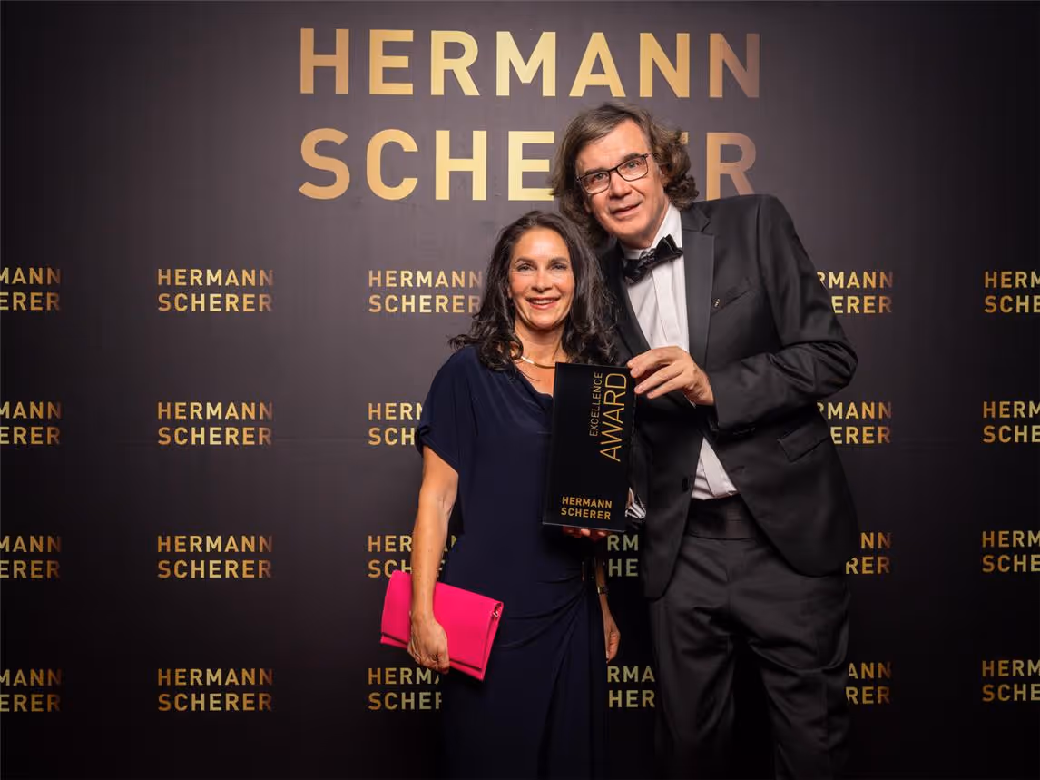 Kathrin Ipse - Expertin für Stressregulation, Nervensystem-Balance und professionelle Hypnose - Ein Mann und eine Frau posieren für ein Foto vor einem schwarzen Hintergrund, auf dem in goldener Schrift der Name „HERMANN SCHERER“ wiederholt ist.