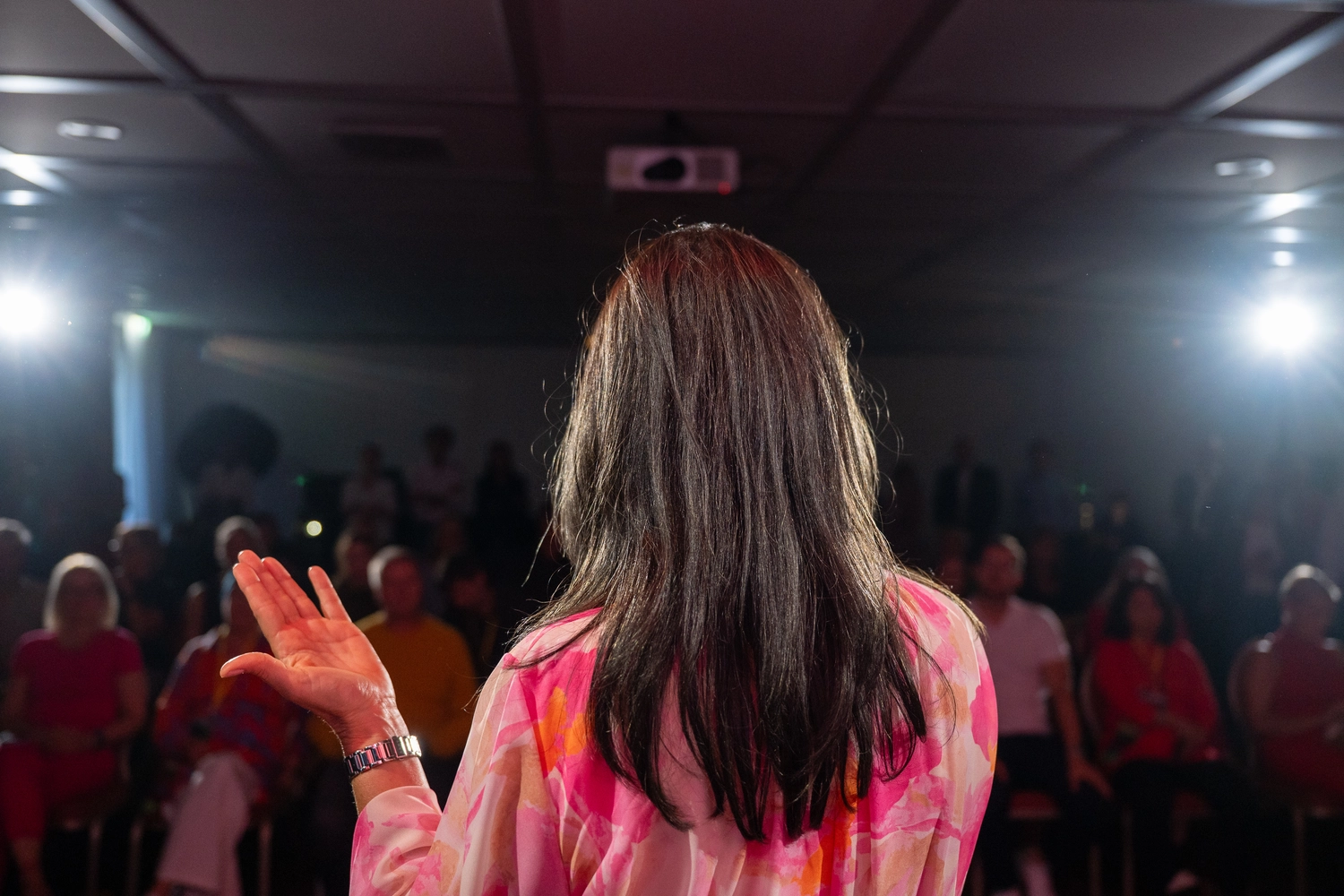 Kathrin Ipse - Expertin für Stressregulation, Nervensystem-Balance und professionelle Hypnose - Eine Person mit langen braunen Haaren steht, von hinten gesehen, auf einer Bühne in einem schwach beleuchteten Raum und blickt zu einem Publikum. Im Hintergrund ist ein Projektor zu sehen.