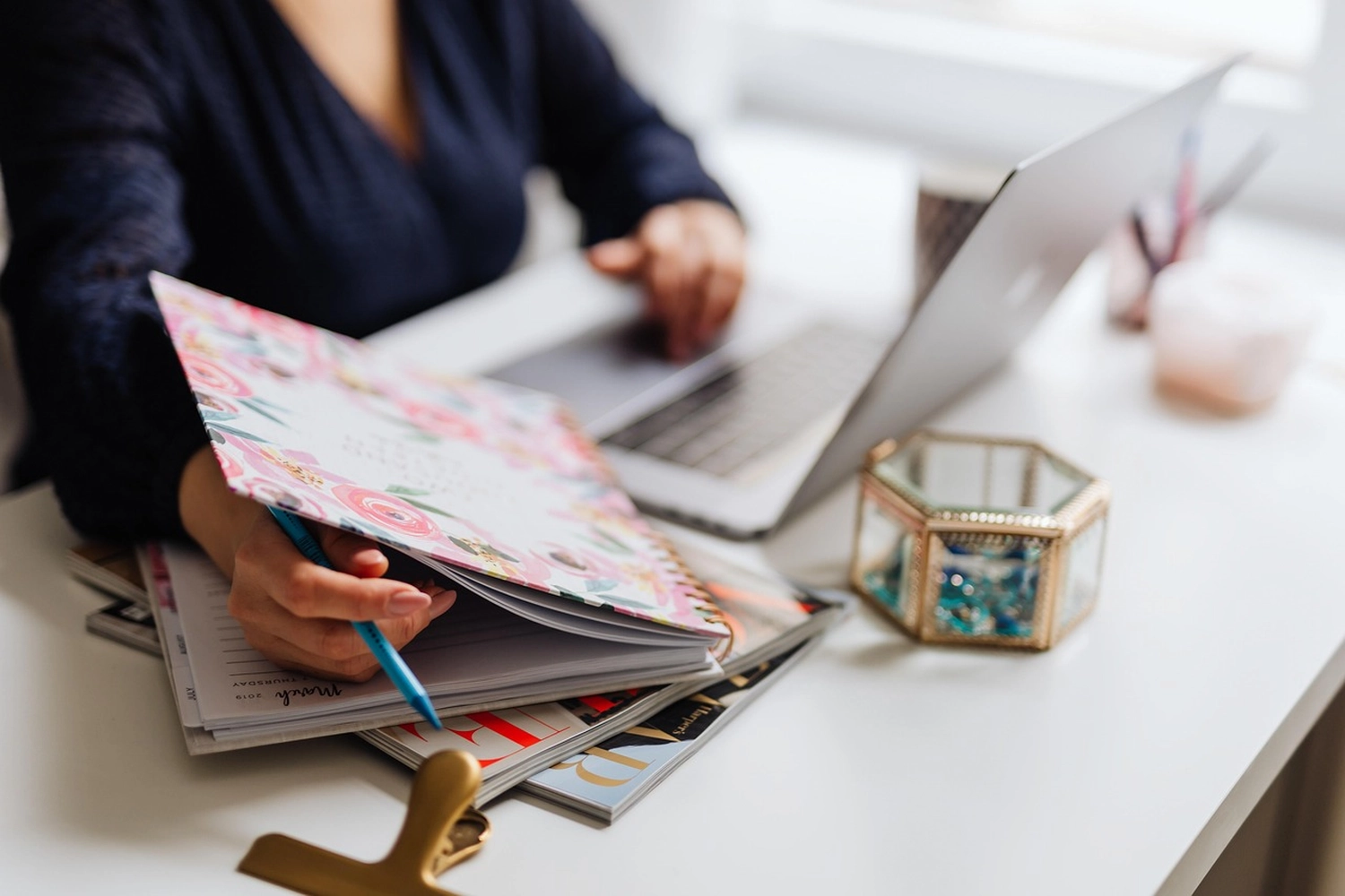 Kathrin Ipse - Expertin für Stressregulation, Nervensystem-Balance und professionelle Hypnose - Die Hände einer Person, die in einem floralen Kalender neben einem Laptop und einer kleinen Glasbox auf einem weißen Schreibtisch schreiben.