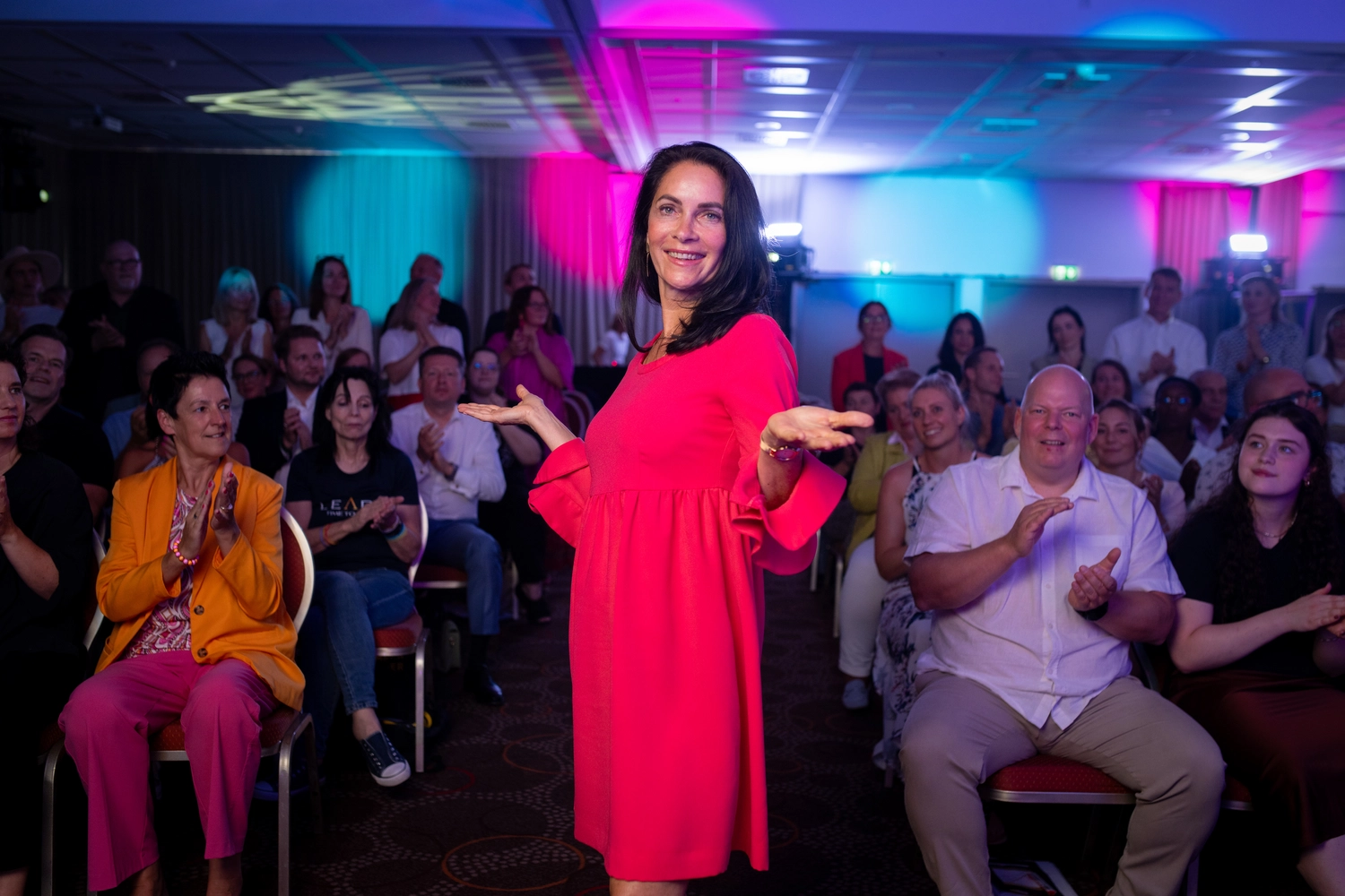 Kathrin Ipse - Expertin für Stressregulation, Nervensystem-Balance und professionelle Hypnose - Eine lächelnde Frau in einem leuchtend pinkfarbenen Kleid steht mit ausgebreiteten Armen auf einer Bühne in einem Konferenzraum und klatscht vor einem Publikum.