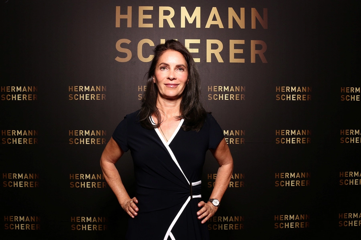 Kathrin Ipse - Expertin für Stressregulation, Nervensystem-Balance und professionelle Hypnose - Eine Frau mit dunklem Haar, die ein marineblaues Wickelkleid mit weißem Besatz trägt, steht vor einer dunklen Wand, die mit sich wiederholendem goldenem Schriftzug bedeckt ist, auf dem „HERMANN SCHERER“ steht.